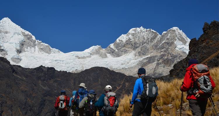 Hikers walking towards snow-covered peaks under a clear blue sky.