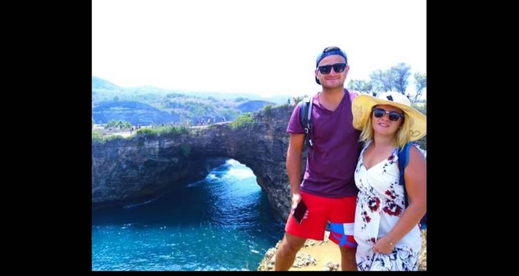 Paar, das in der Nähe eines natürlichen Felsenbogens steht, mit blauem Meer im Hintergrund.