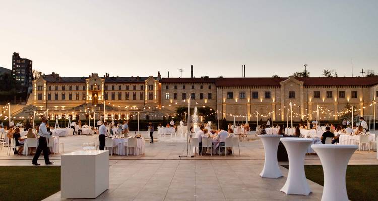 Evento al aire libre con personas cenando frente a un edificio grandioso, bajo luces colgantes.
