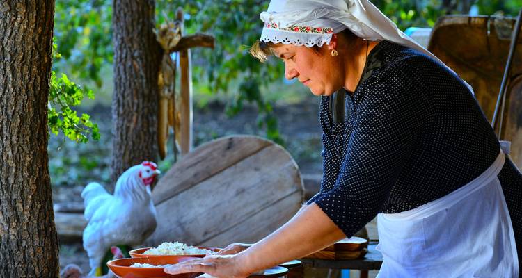 Mujer con cofia preparando una comida al aire libre con una gallina cerca.