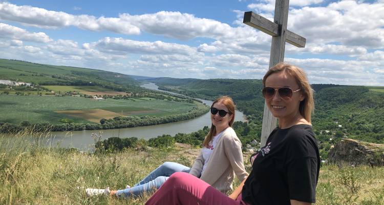 Dos mujeres sentadas en la cima de una colina junto a una cruz, contemplando un río y un valle.