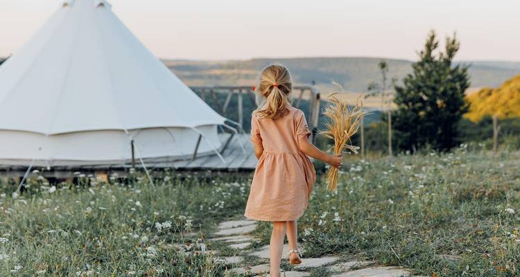 Niña caminando hacia una tienda de campaña en un campo, sosteniendo trigo en su mano.