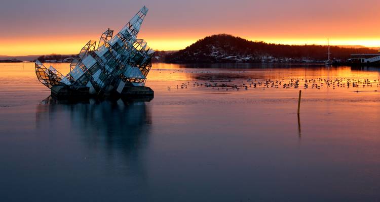 Abstract sculpture partially submerged in water with a sunset backdrop.