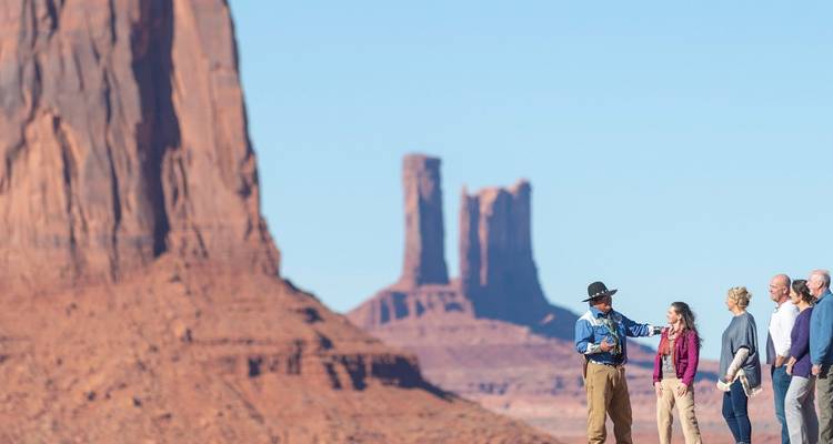 Navajo-Führer gestikuliert, während Besucher vor den hoch aufragenden Sandsteinbergen des Monument Valley stehen.