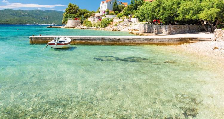 Mer turquoise transparente et plage de galets tranquille avec petit bateau amarré près des maisons de l'île croate.