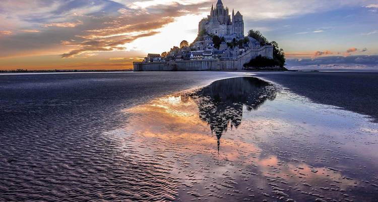 Mont Saint-Michel se eleva sobre las llanuras de marea reflectantes bajo un cielo dramático del amanecer.