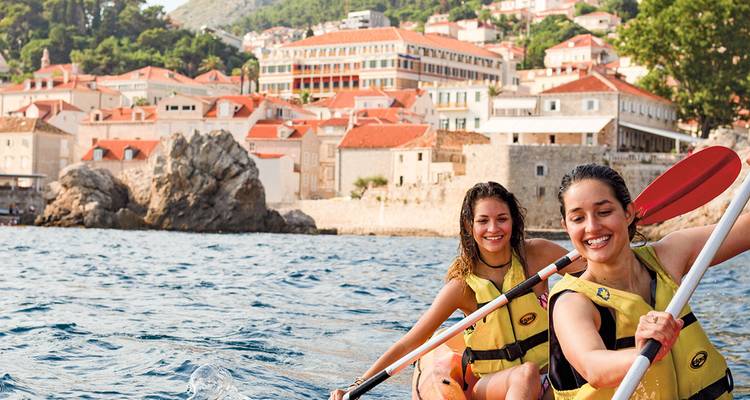 Dos mujeres navegando en kayak en las aguas azules del Adriático con los tejados de terracota de Dubrovnik al fondo.