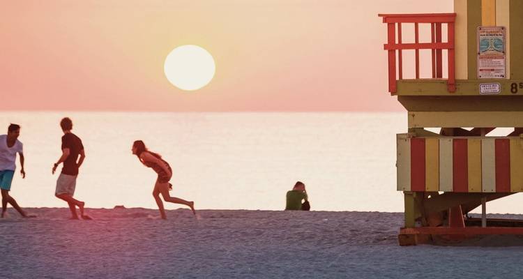 Silhouettenhafte Strandbesucher bei Sonnenuntergang mit einem Rettungsschwimmerturm und glühender orangefarbener Sonne über ruhigem Wasser.