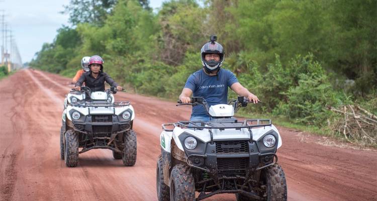 Drei Reisende fahren mit Quads eine rote Schotterstraße entlang, die von üppiger grüner Vegetation gesäumt wird.