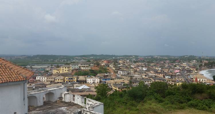 Erhöhte Aussicht auf eine Küstenstadt in Ghana mit sanft gewellten grünen Hügeln und dicht gedrängten Clustern von niedriggeschossigen Gebäuden.
