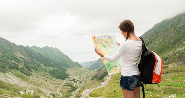 Une randonneuse étudie une carte de la Roumanie surplombant la route sinueuse du Transfăgărășan et la vallée verdoyante.
