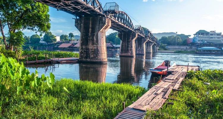 Le pont historique en acier enjambe un fleuve tranquille, flanqué de végétation luxuriante et d'une jetée en bois avec un bateau à longue queue.
