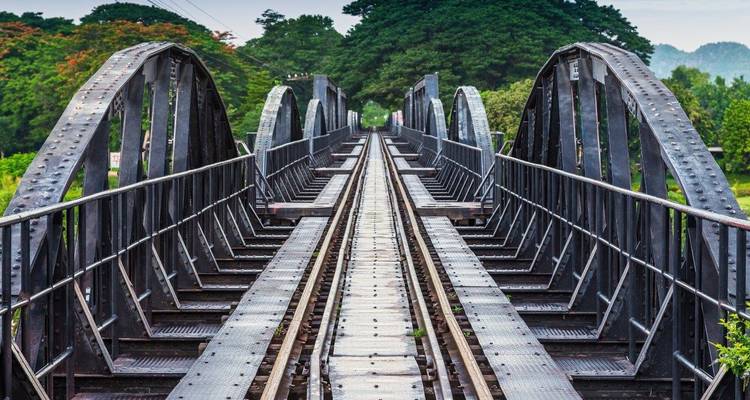 Vue symétrique le long des voies du pont en acier noir iconique sur la rivière Kwaï disparaissant dans une végétation luxuriante.