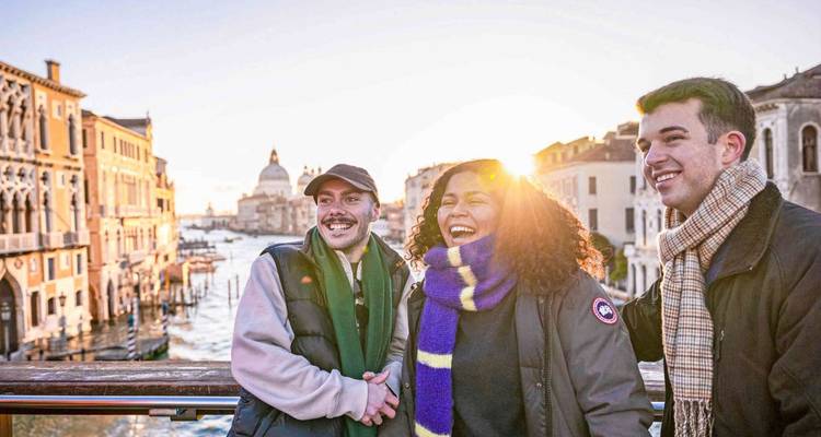 Three smiling friends stand on a bridge over Venice's Grand Canal as the sun sets behind historic palaces and domes.