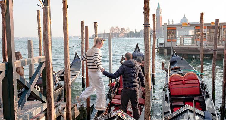 Viajeros abordando góndolas en un muelle de madera con el horizonte de Venecia al fondo.