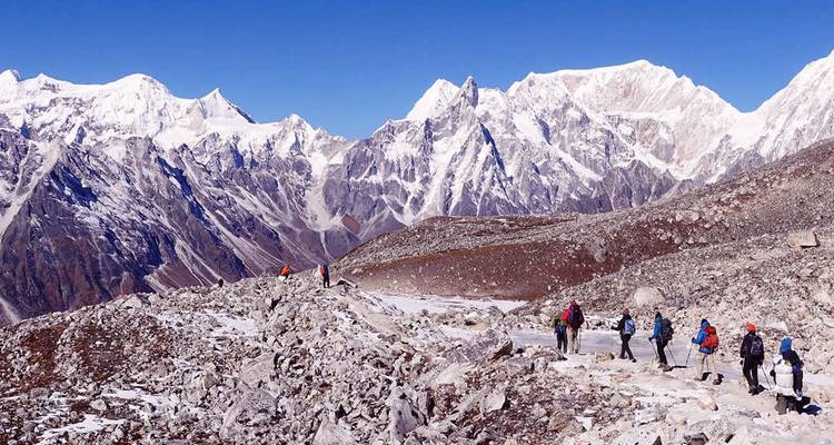 Randonneurs traversant un sentier de montagne rocailleux avec des sommets enneigés.