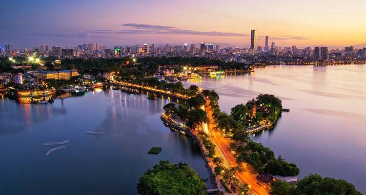 Twilight aerial of Hanoi’s West Lake with illuminated roads curving into the glowing city skyline