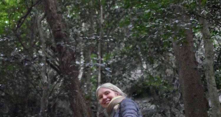 Frau lächelt, während sie auf einem dämmrigen Waldweg steht, umgeben von dunklem Laub.