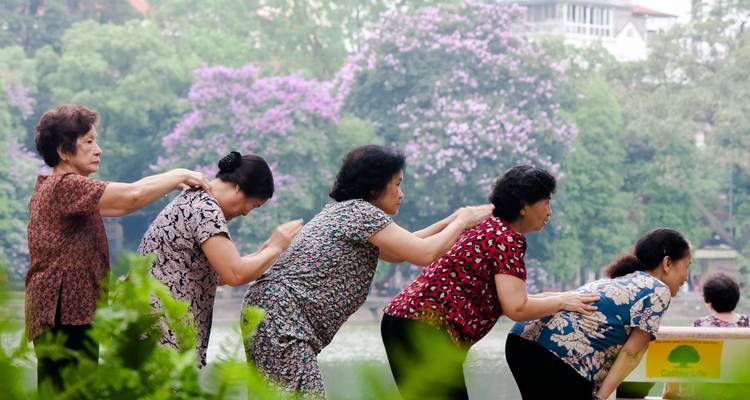 Seis mujeres mayores están de pie en fila junto a un lago, dándose masajes en los hombros entre árboles frondosos con flores moradas.