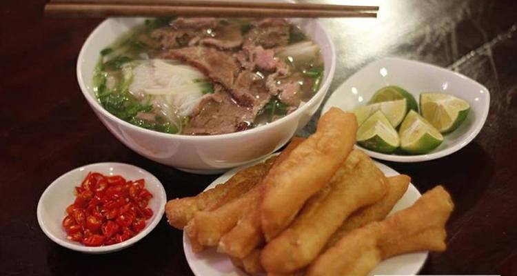 Un tazón humeante de pho vietnamita con carne, servido con gajos de lima, chiles en rodajas y palitos de masa frita crujientes.