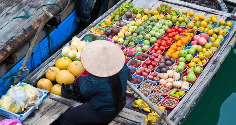 An overhead view of a vendor in a conical hat selling a colorful array of fruits from a wooden boat at a floating market.
