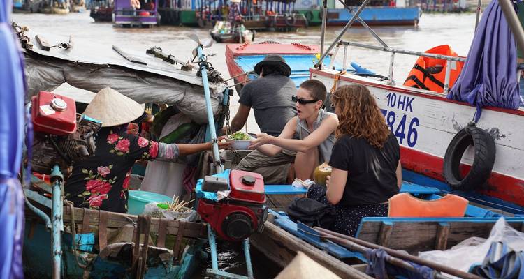 Tourists in wooden boats buy food from a vendor at a busy Mekong floating market.