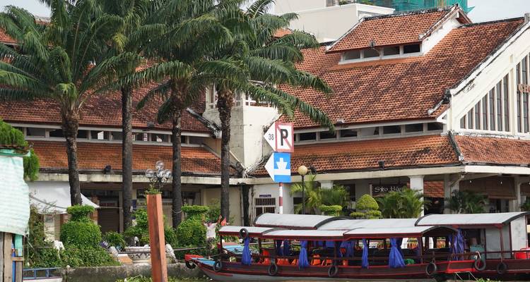 Colonial-era riverside building with red-tiled roof and palm trees behind a moored boat.