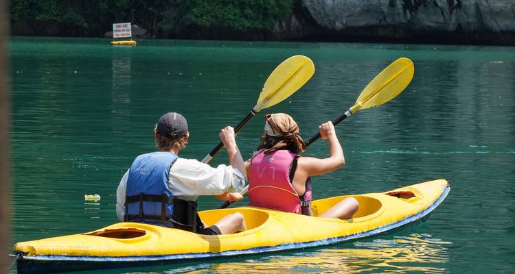 Dos viajeros reman en un kayak amarillo brillante a través de aguas esmeralda tranquilas bajo acantilados.