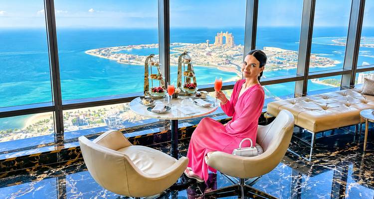 Femme élégante dégustant un high-tea avec vue panoramique sur la Paume de Jumeirah et le Golfe Arabique depuis un salon situé dans un gratte-ciel.