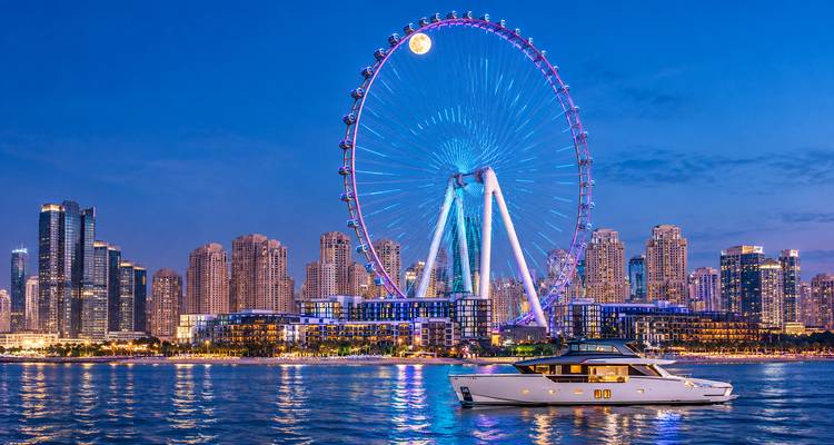 Vue nocturne de l'horizon de Dubai Marina avec la grande roue Ain Dubai illuminée et un yacht de luxe sur des eaux calmes.
