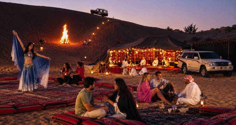 Camp du désert en soirée avec tapis, lanternes et clients regardant une danseuse du ventre près d'un feu de joie flamboyant sous les cieux crépusculaires.