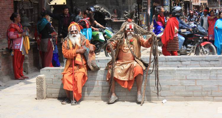 Zwei farbenfroh gekleidete Sadhus ruhen auf einer Steinbank, während das geschäftige Treiben der Straße hinter ihnen vorbeizieht.