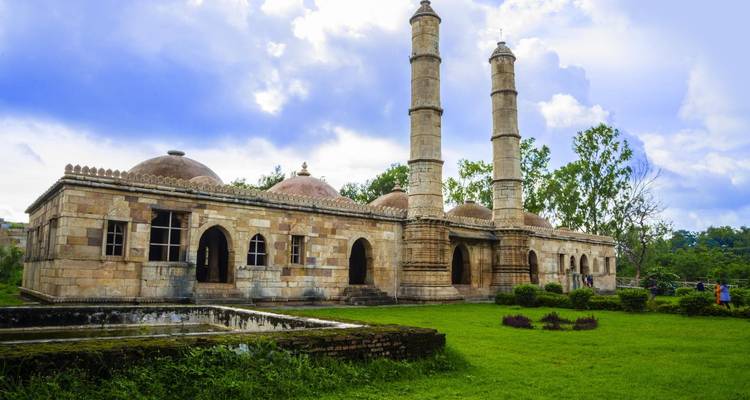 Complejo histórico de mezquita de piedra con dos minaretes y jardines bien cuidados bajo nubes dramáticas.
