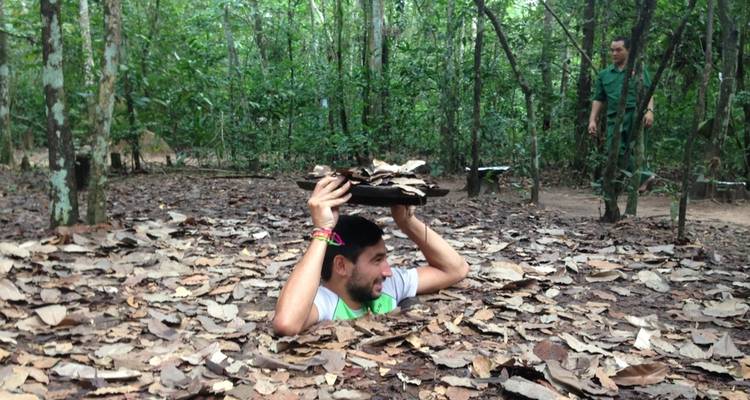 Person, die aus einem Vietcong-Tunnel mit Blättern obendrauf auftaucht, und eine andere Person in der Nähe im Wald.