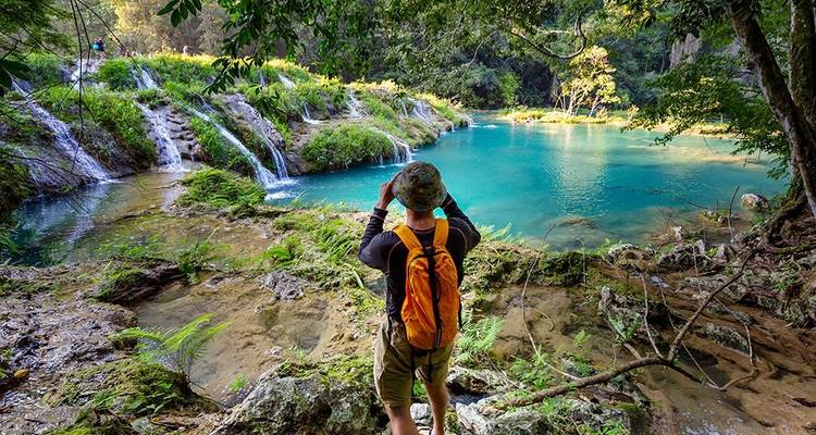 Wandelaar met oranje rugzak fotografeert levendige turquoise poelen en kleine watervallen in dichte jungle.