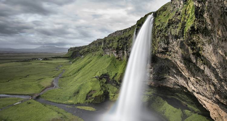 Wasserfall, der über moosbewachsene Klippen in eine grasige Landschaft mit bewölktem Himmel hinabstürzt.