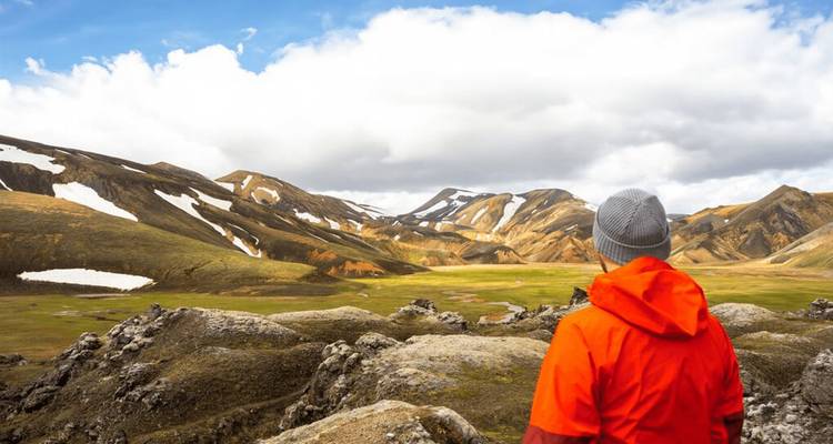 Viajero con una chaqueta naranja brillante de pie en rocas de lava mirando hacia colinas de riolita coloridas y parches de nieve en Islandia