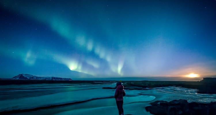 Un voyageur solitaire observe l'aurore scintiller au-dessus d'une rivière gelée et de collines lointaines dans le crépuscule profond.