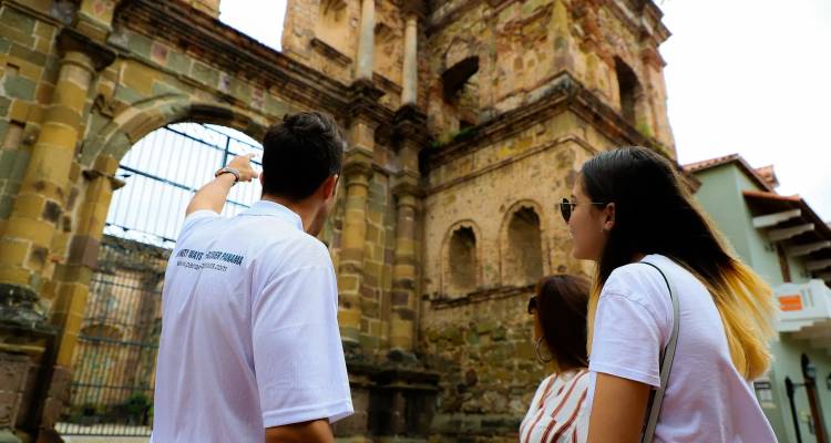 Los visitantes escuchan a un guía que señala la fachada de una antigua iglesia de piedra a través de una puerta de hierro forjado.