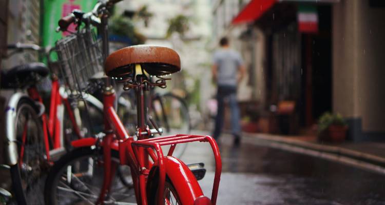 Una bicicleta roja brillante está de pie en un callejón lluvioso con una figura borrosa caminando alejándose en desenfoque suave.