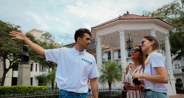 Un guía hace gestos en una plaza frondosa cerca de un gazebo blanco mientras dos visitantes lo escuchan con interés.