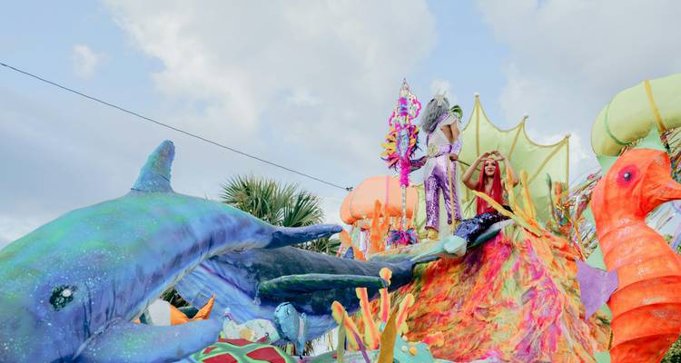 Carroza de carnaval colorida con criaturas marinas y artistas en trajes brillantes bajo un cielo azul.