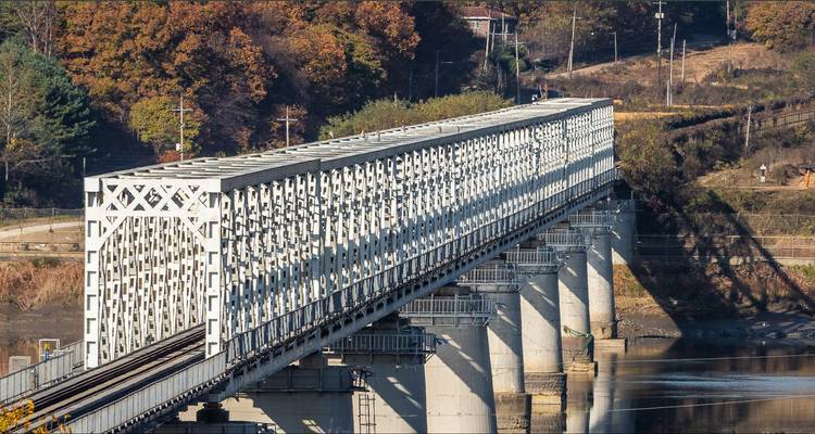 Long pont ferroviaire à treillis blanc enjambant une rivière calme avec des arbres d'automne en arrière-plan.