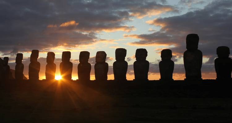 Línea de estatuas moái recortadas contra el horizonte mientras sale el sol, proyectando rayos dorados a través de nubes dramáticas.