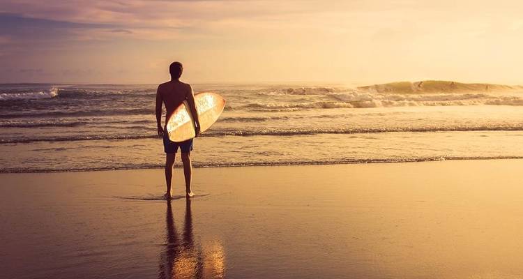 Surfeur debout sur une plage, regardant l'océan pendant le coucher de soleil.