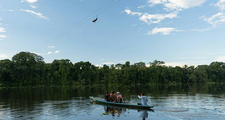 Des gens dans un bateau sur une rivière avec de la verdure et quelqu'un qui fait de la tyrolienne au-dessus.