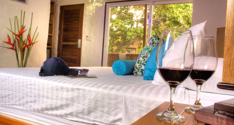 Acogedora habitación de hotel con sábanas blancas impecables, cojines de colores y dos copas de vino tinto en la mesita de noche.