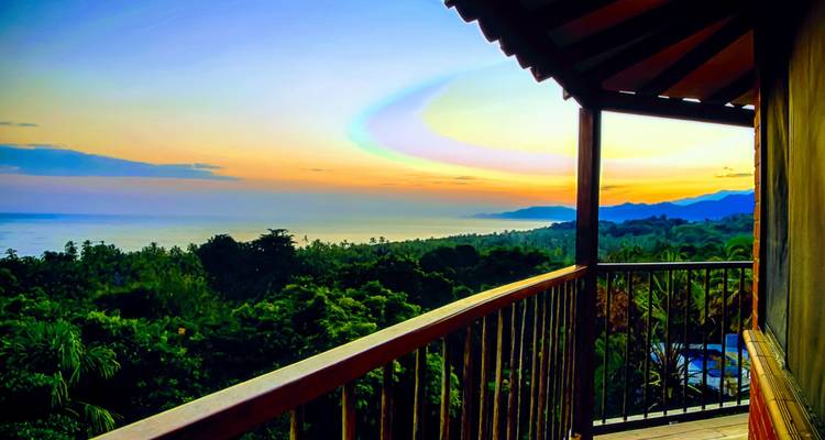 Un colorido balcón con vistas al dosel de la selva tropical y al mar distante durante una puesta de sol vibrante.