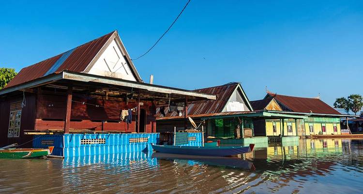 Maisons en bois aux couleurs vives d'un village flottant se reflétant dans des eaux calmes sous un ciel bleu dégagé.