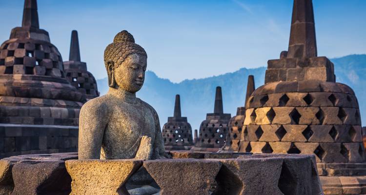 Statue de Bouddha en pierre trône sereinement parmi les stupas en forme de cloche de Borobudur au lever du soleil avec des montagnes brumeuses en arrière-plan.